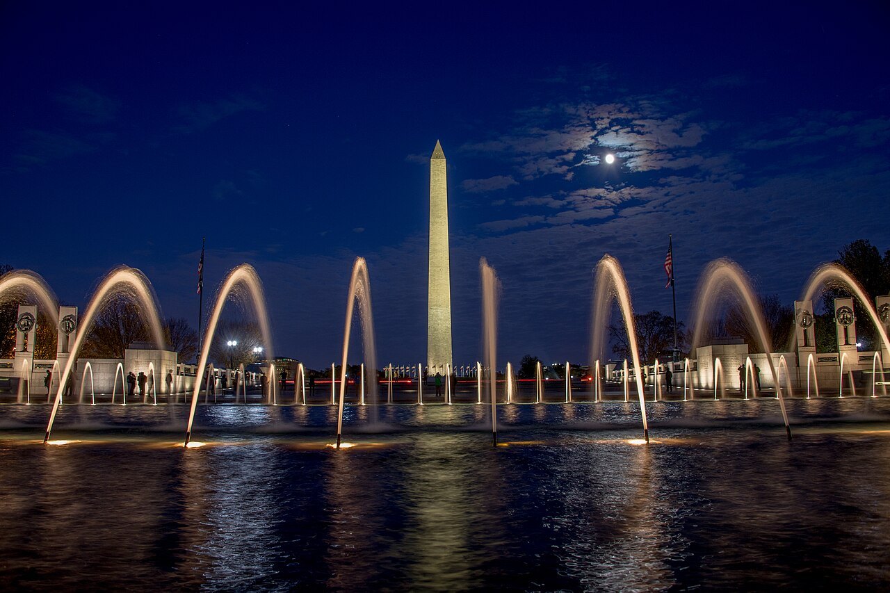 The Washington Monument by moonlight beyond the lit fountains of the World War II Memorial
